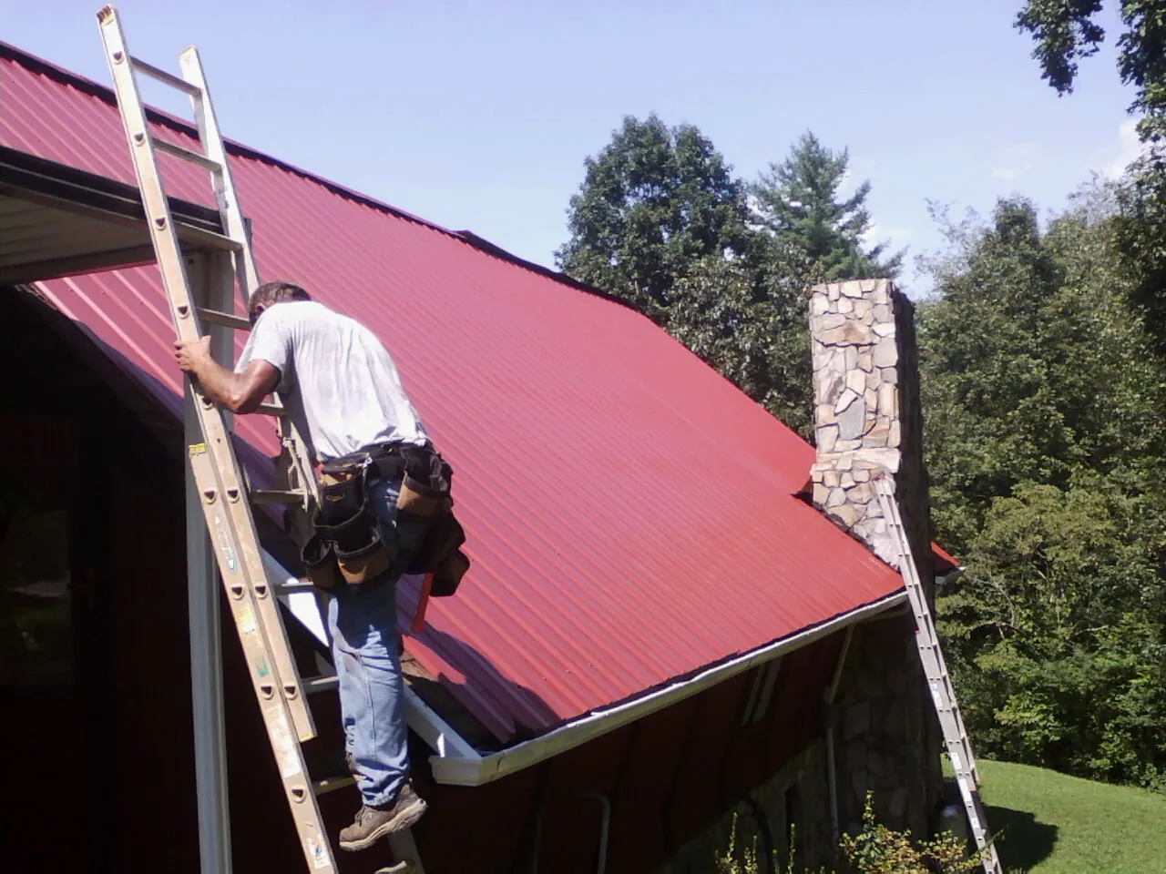Roofer on a ladder beside a red metal roof and stone chimney — Soffit & Fascia Repair by Roofing Brothers in Lufkin